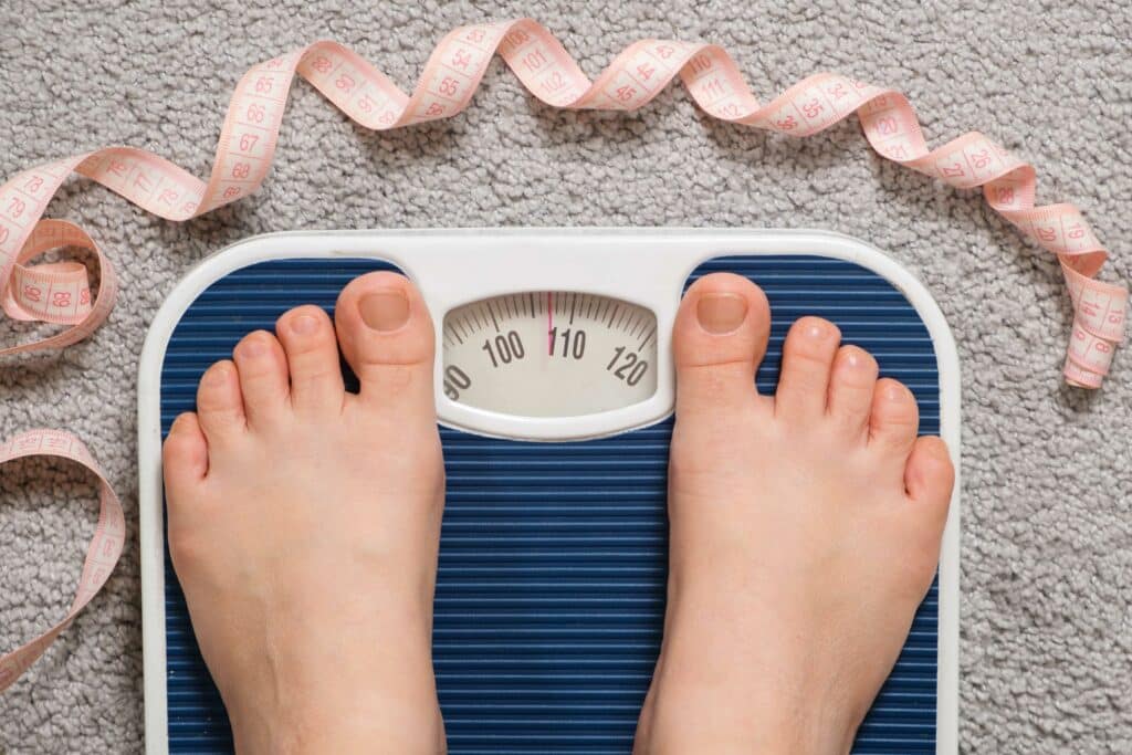 Women's bare feet on floor scales and measuring tape, weight 100-110 kilograms, top view. The idea of obesity, weight loss and excess weight