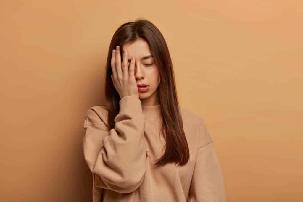 I cannot look at this mess. Frustrated tired woman makes facepalm, stands displeased and uninterested, sighs from tiredness after working long time, wears sweatshirt, isolated on brown wall