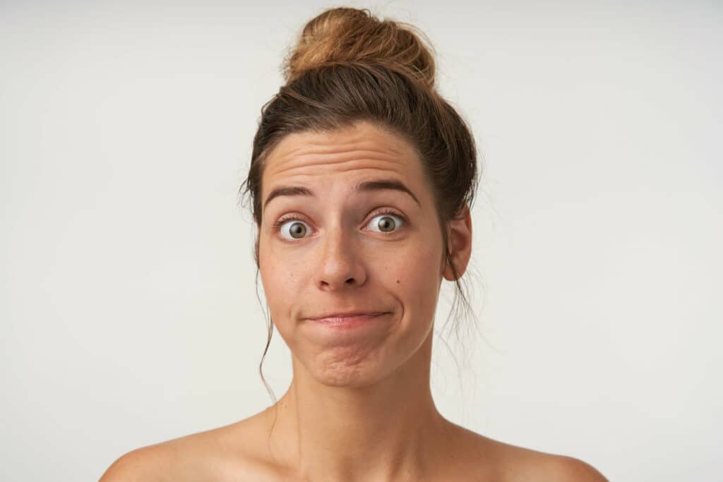 Close-up of attractive young woman posing over white background with bewildered face, wearing bun hairstyle and no make-up, wrinkling forehead and pursing lips