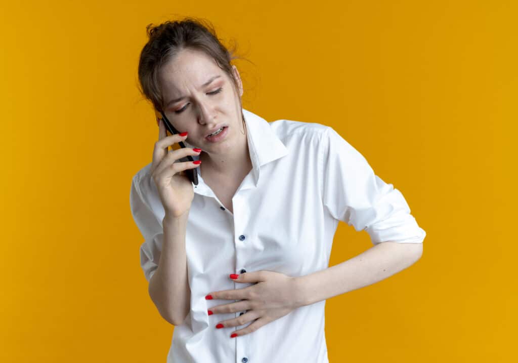 young aching blonde russian girl puts hand on belly talking on phone isolated on orange background with copy space