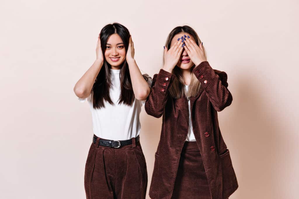 Dark-haired girl smiles and covers her ears, blonde woman in corduroy suit covers her eyes with her hands